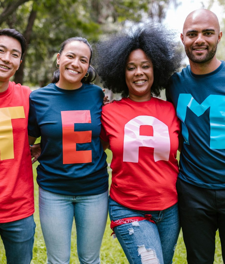A diverse group of adults bonding and smiling in a park wearing TEAM shirts.